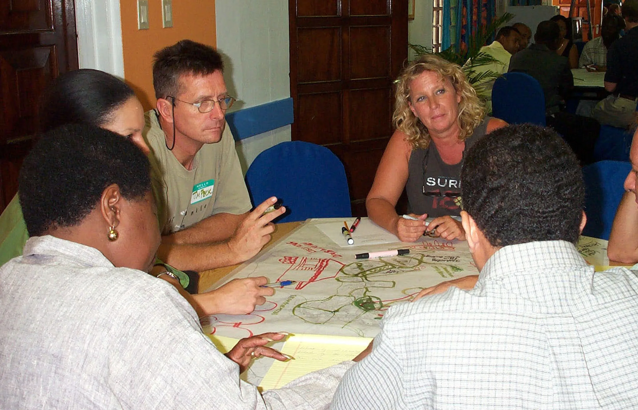 Tim Peck and residents of Road Town around a table, drawing over a plan of the town