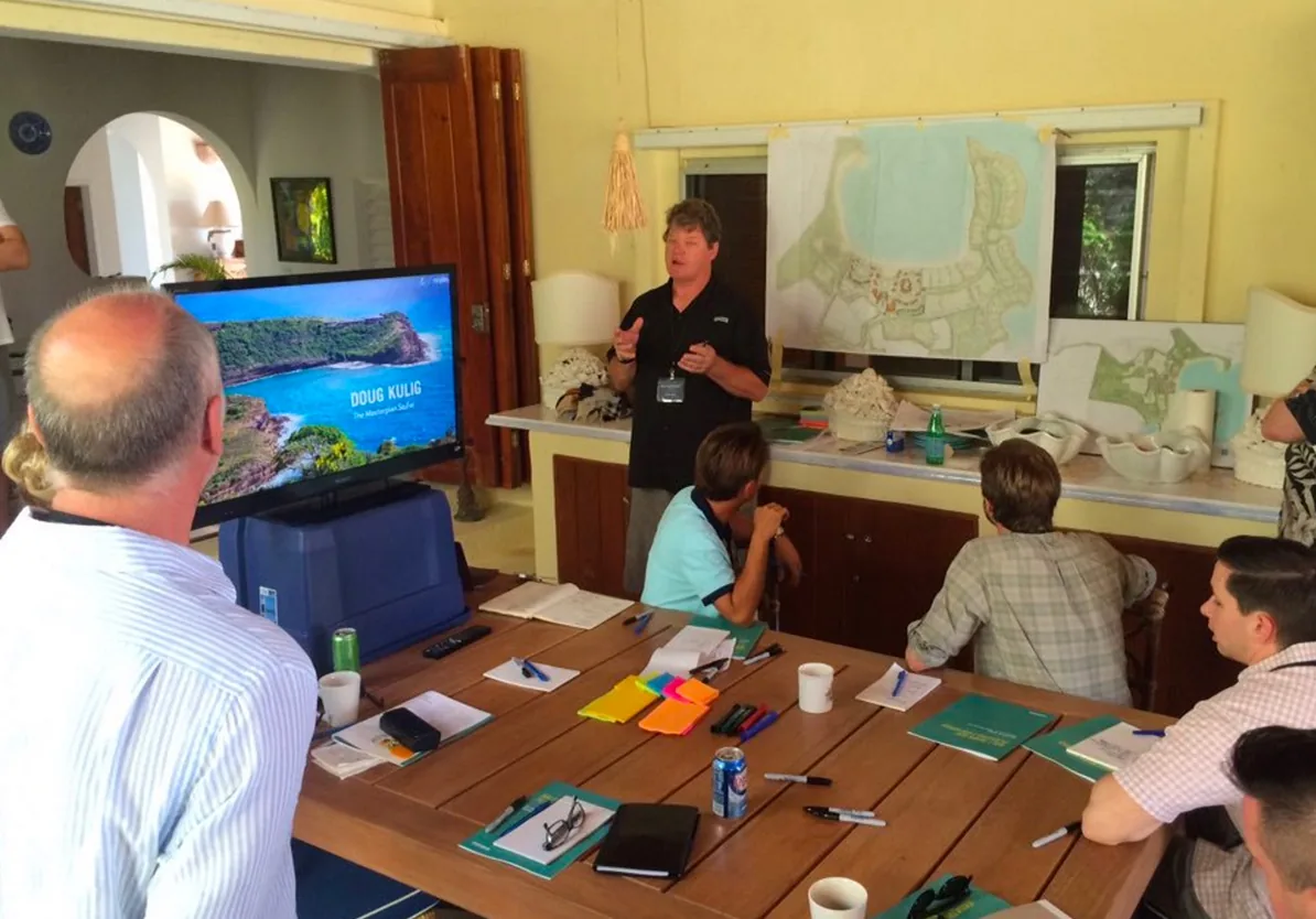 Doug Kulig presenting a masterplan to a team around a table in 2008, a screen and pinned drawings behind him