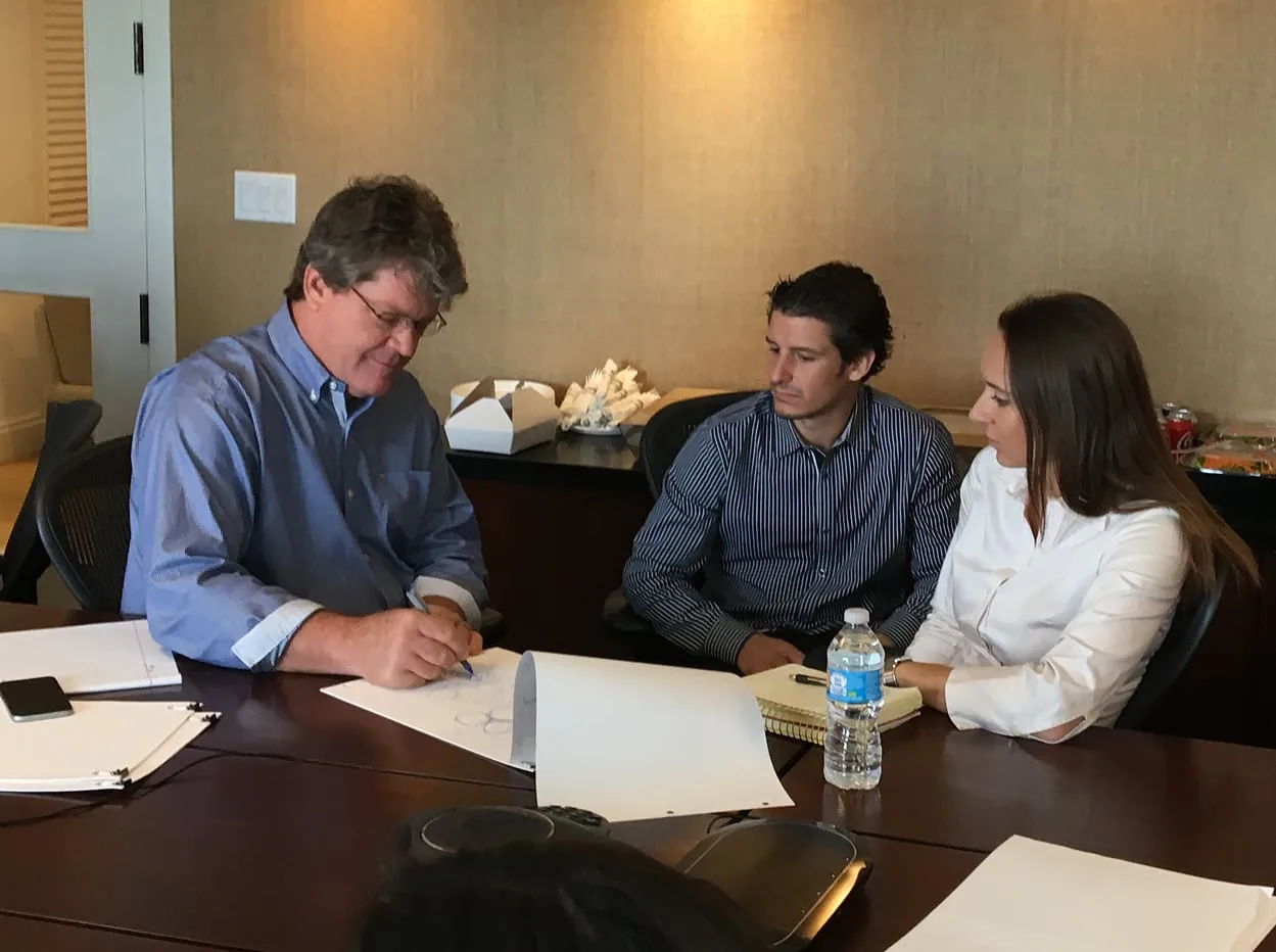 Doug Kulig writing at a boardroom table, two younger colleagues beside him