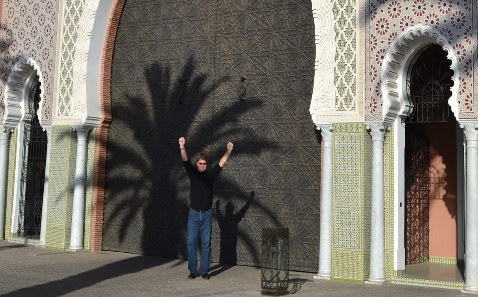 Doug Kulig with both arms raised in front of the great bronze doors of the Royal Mansour, a palm's shadow across them
