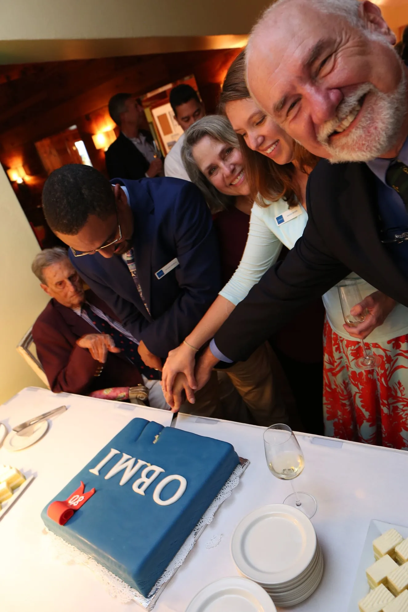 The OBMI team cutting a blue cake marked eighty, their hands stacked on the knife