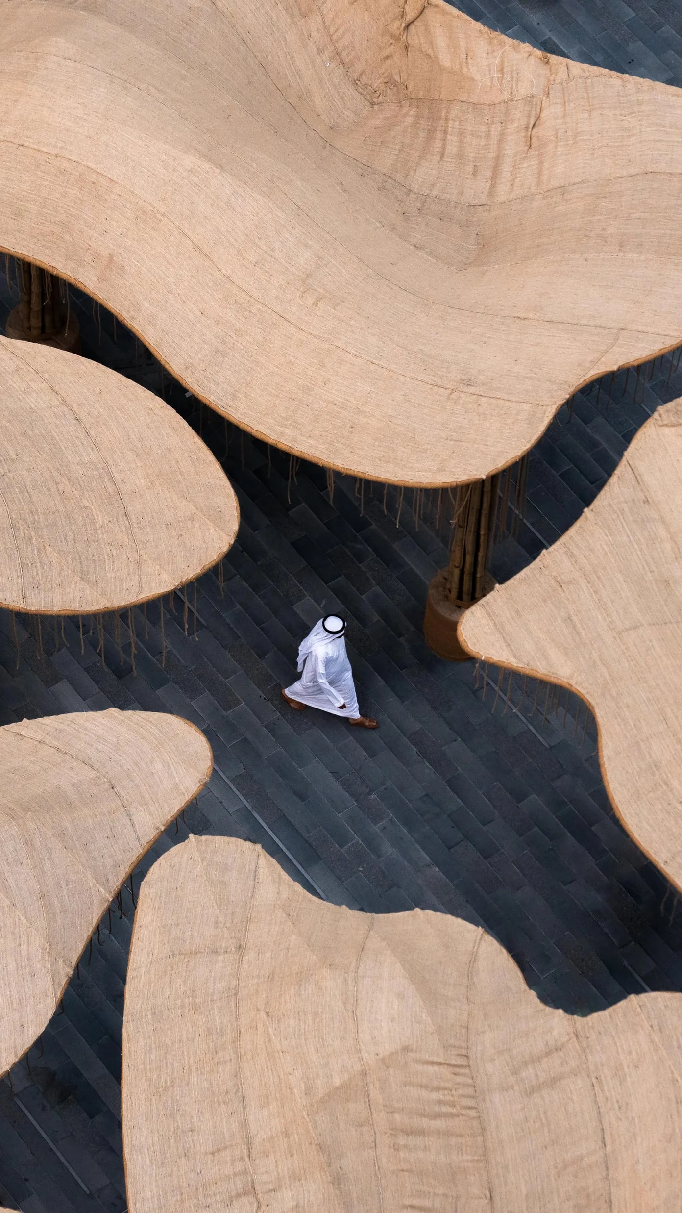 The pavilion from above: undulating jute canopies over a paved square, one man in white walking between them