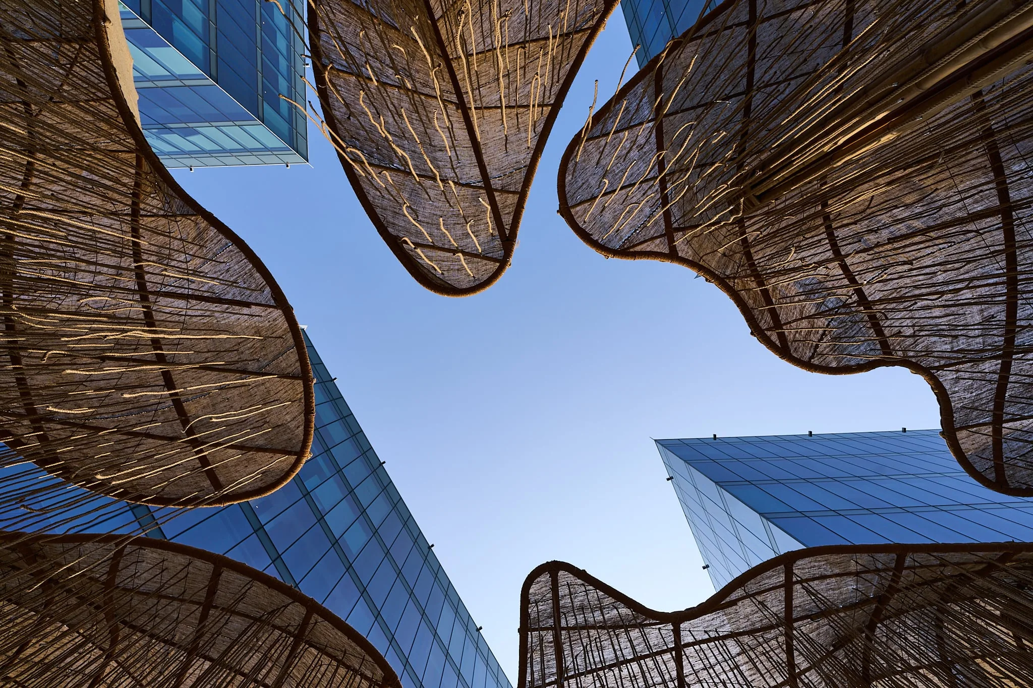 The pavilion's jute canopies seen from beneath, glass towers and blue sky between them