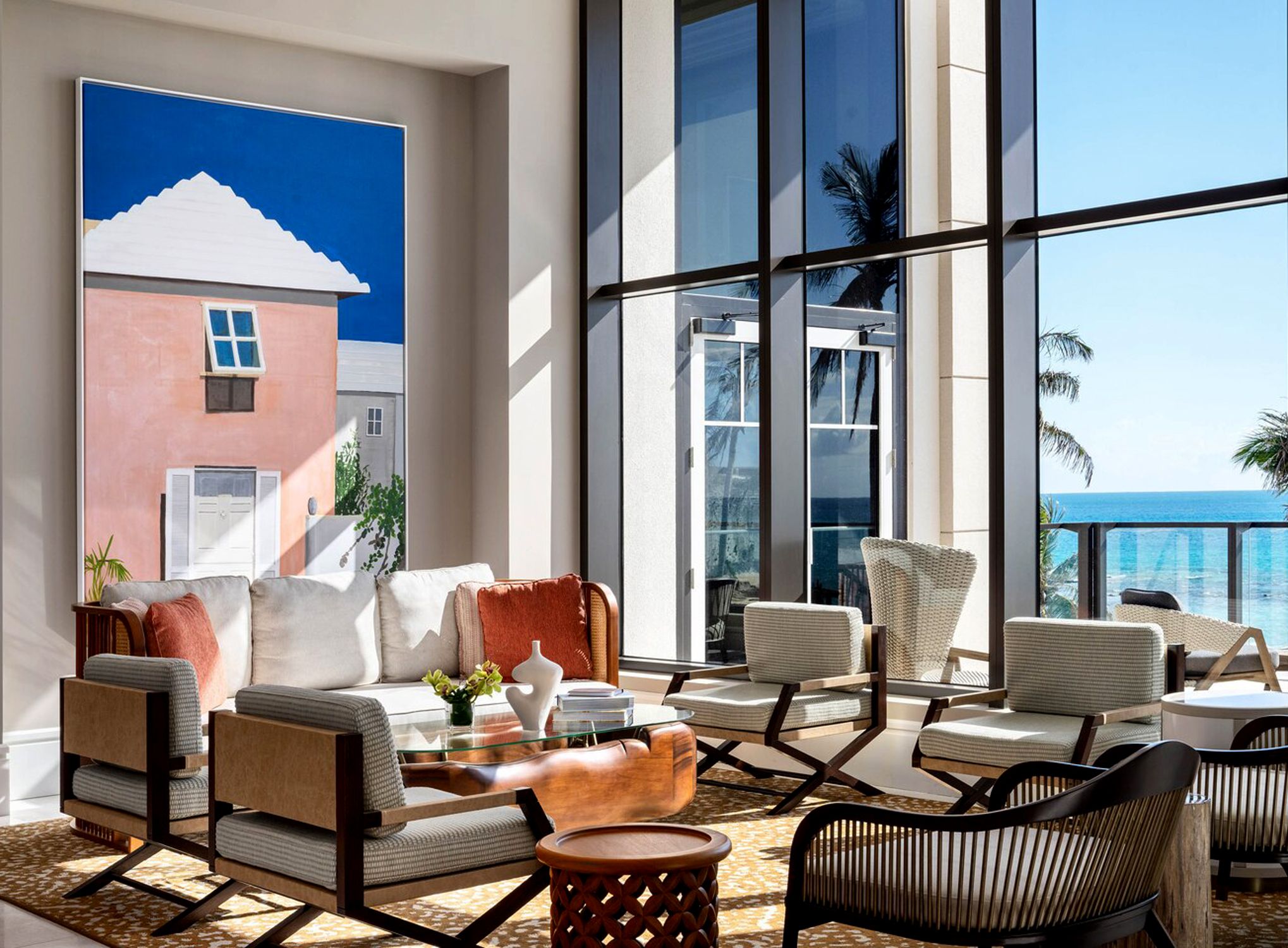 A lounge with a painting of a pink Bermuda house, cane chairs and coral cushions