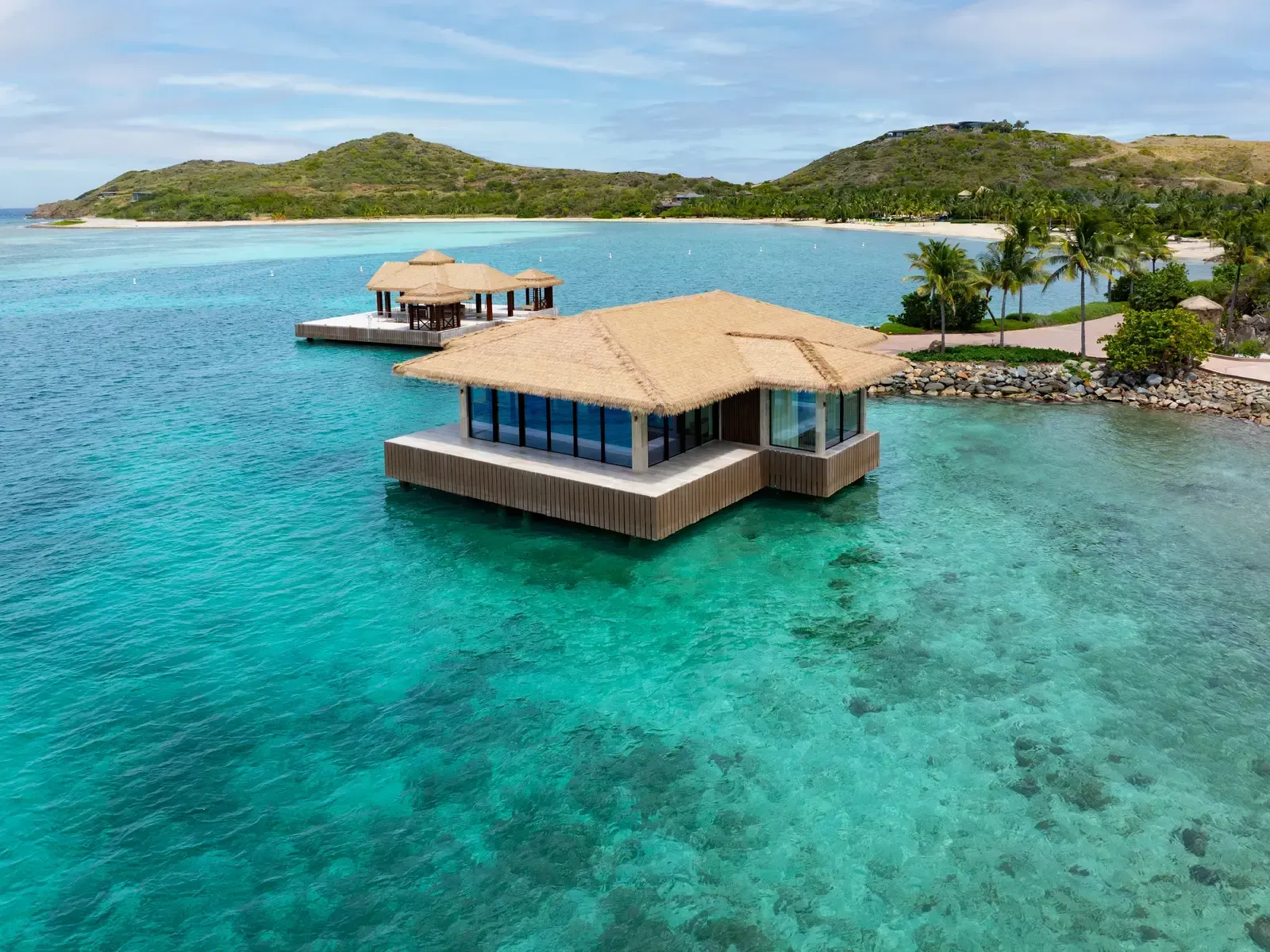 The pavilion from above, the bay, a beach and palms behind it, green hills beyond