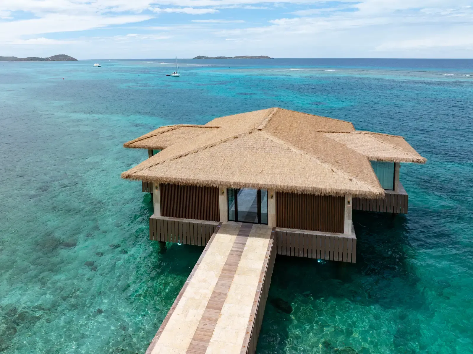 A thatched pavilion over turquoise water, a jetty leading to it, islands on the horizon