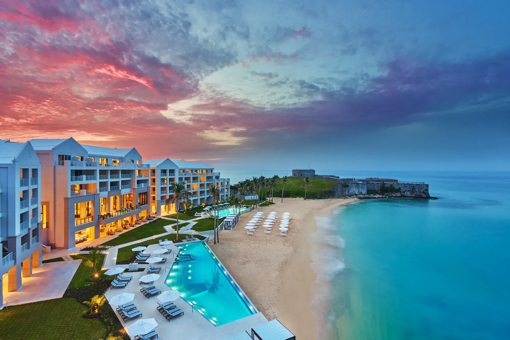 The hotel at sunset from above, its pool lit, the beach curving to Fort St Catherine