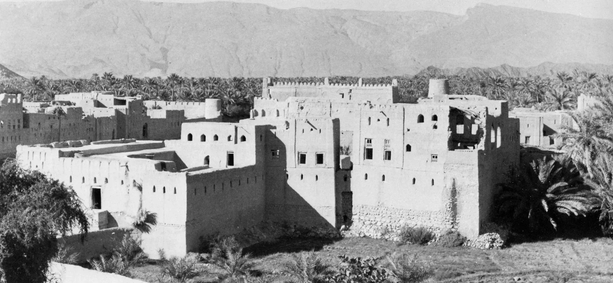 Early photograph of a walled Omani town among date palms, mountains behind