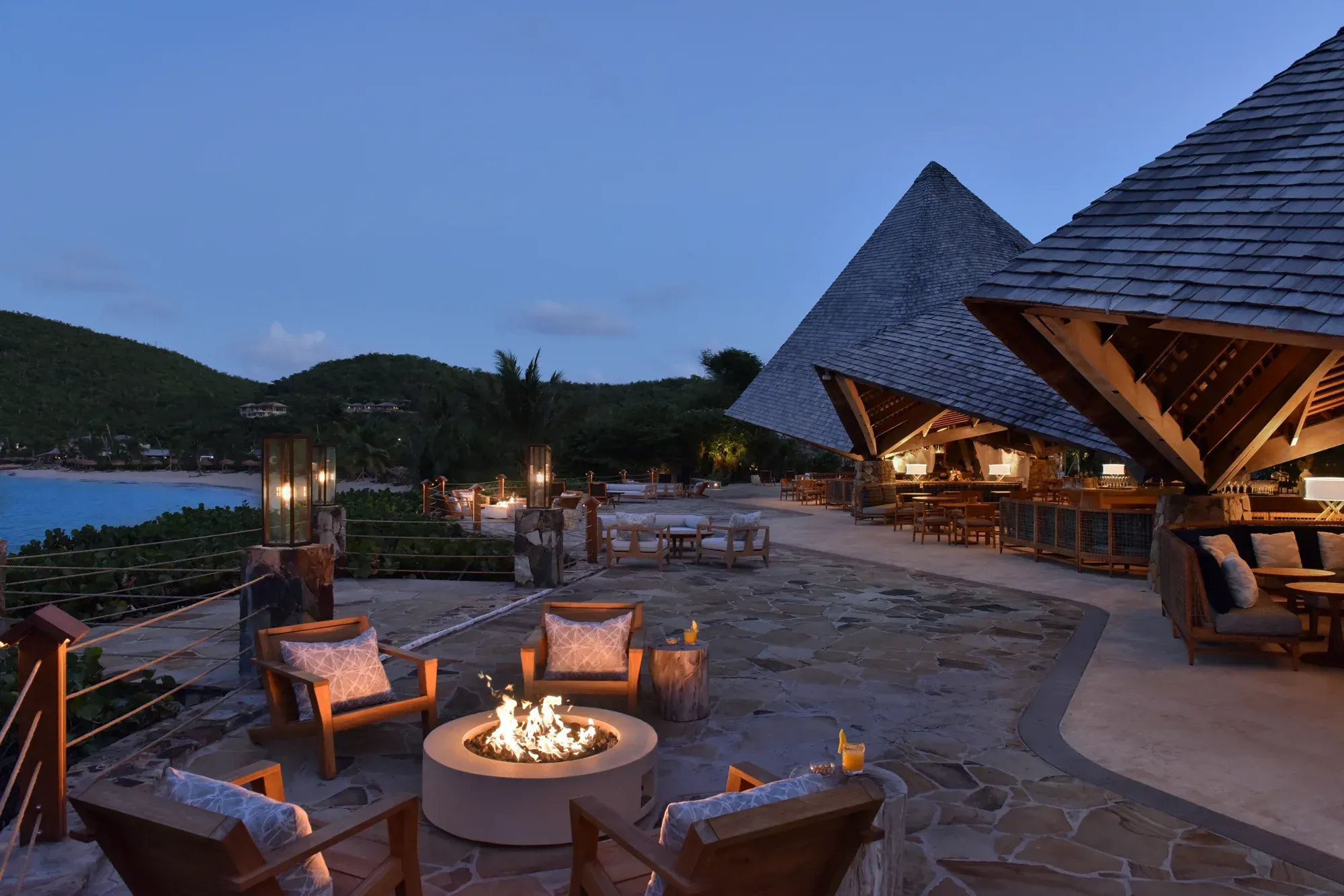 A stone terrace at dusk, armchairs around a fire pit, the pyramid roofs lit behind and the bay beyond