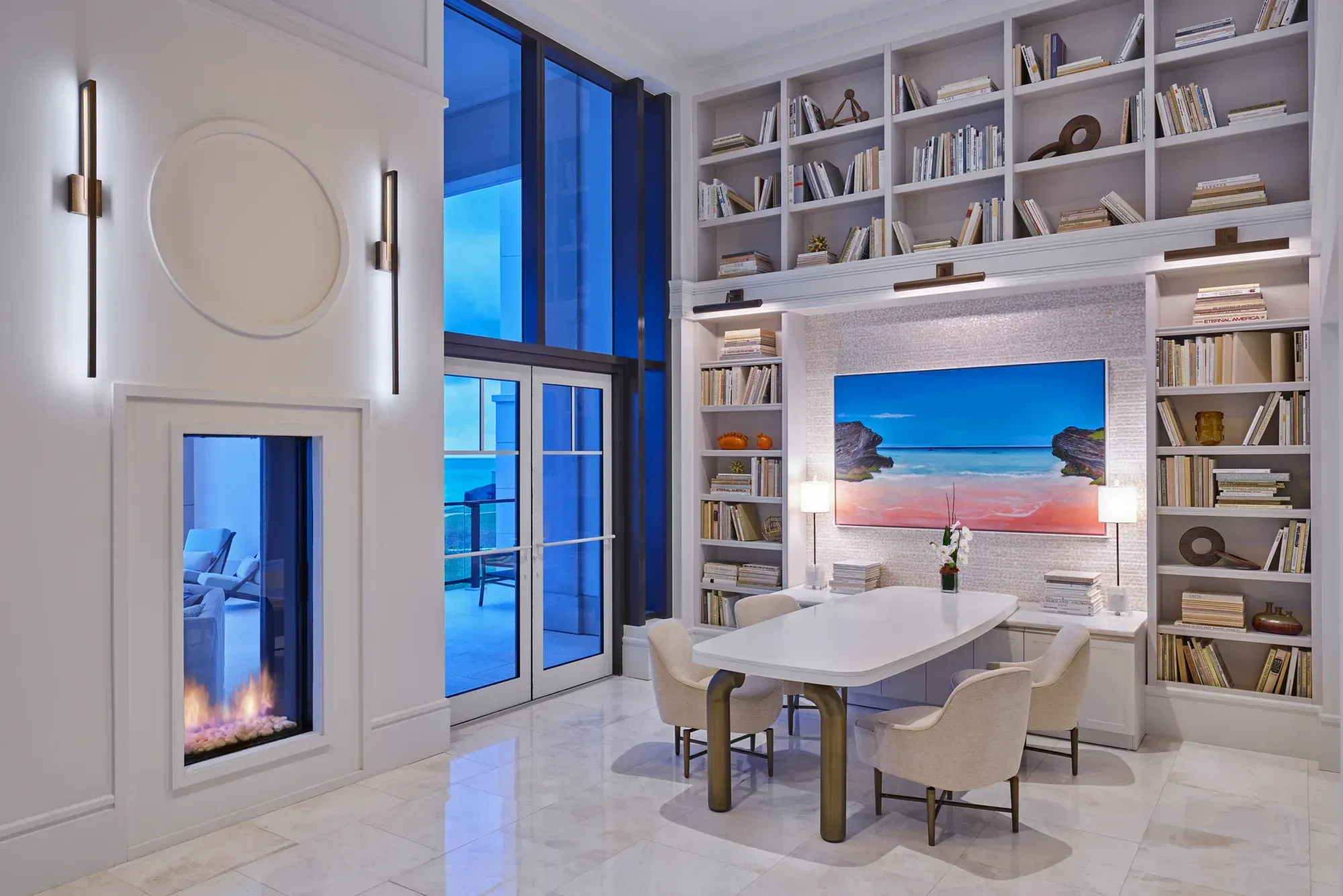 A library at dusk: a fireplace, shelves of books and a painting of the pink beach
