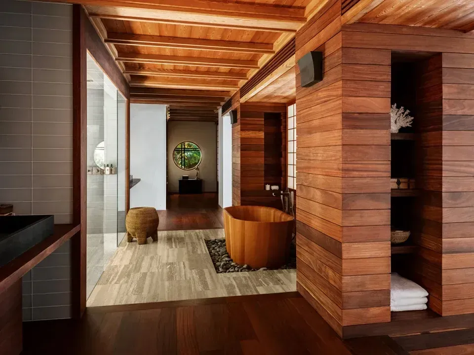 A timber soaking tub on river stones in a bathroom of cedar, a round window beyond