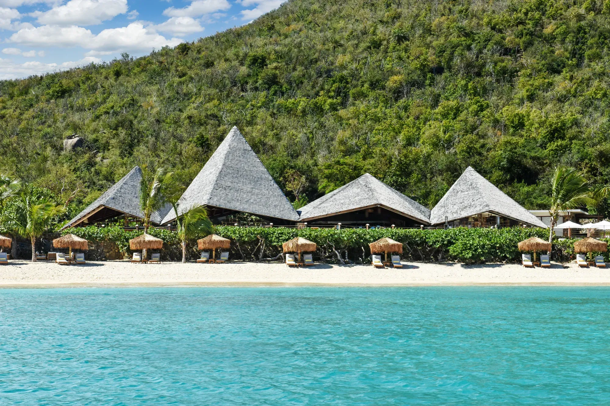 The pavilions along the beach in daylight, thatched parasols on the sand, the hill behind