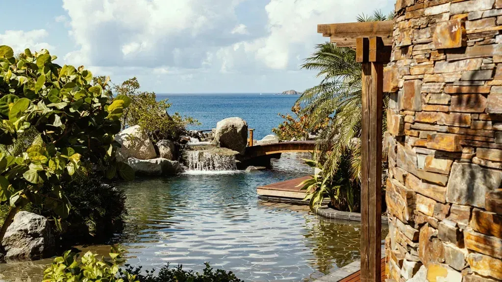 A stone wall beside a pond, a waterfall and a timber bridge, the channel beyond