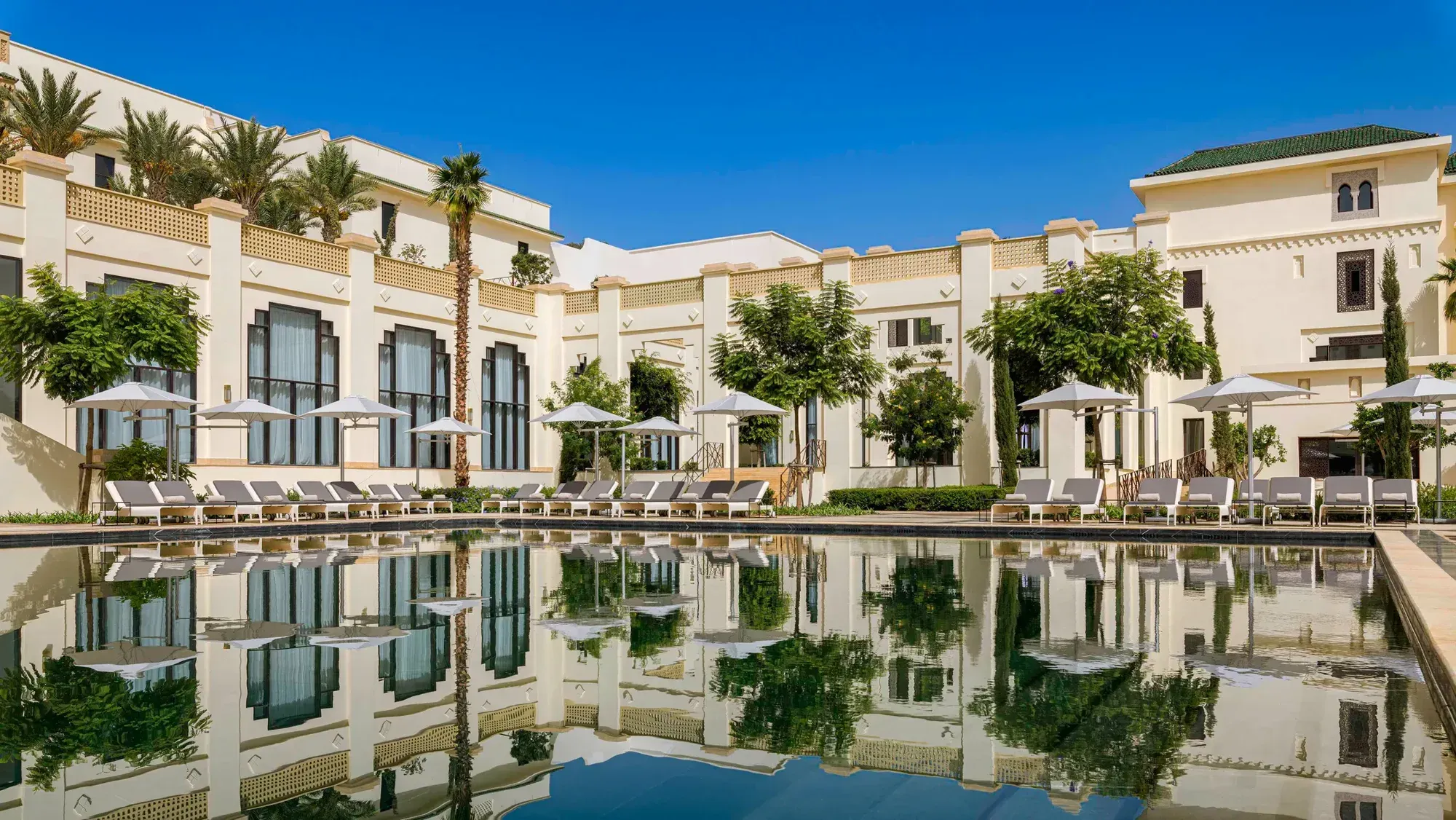 The pool before a white facade, cypresses and palms, parasols along its edge