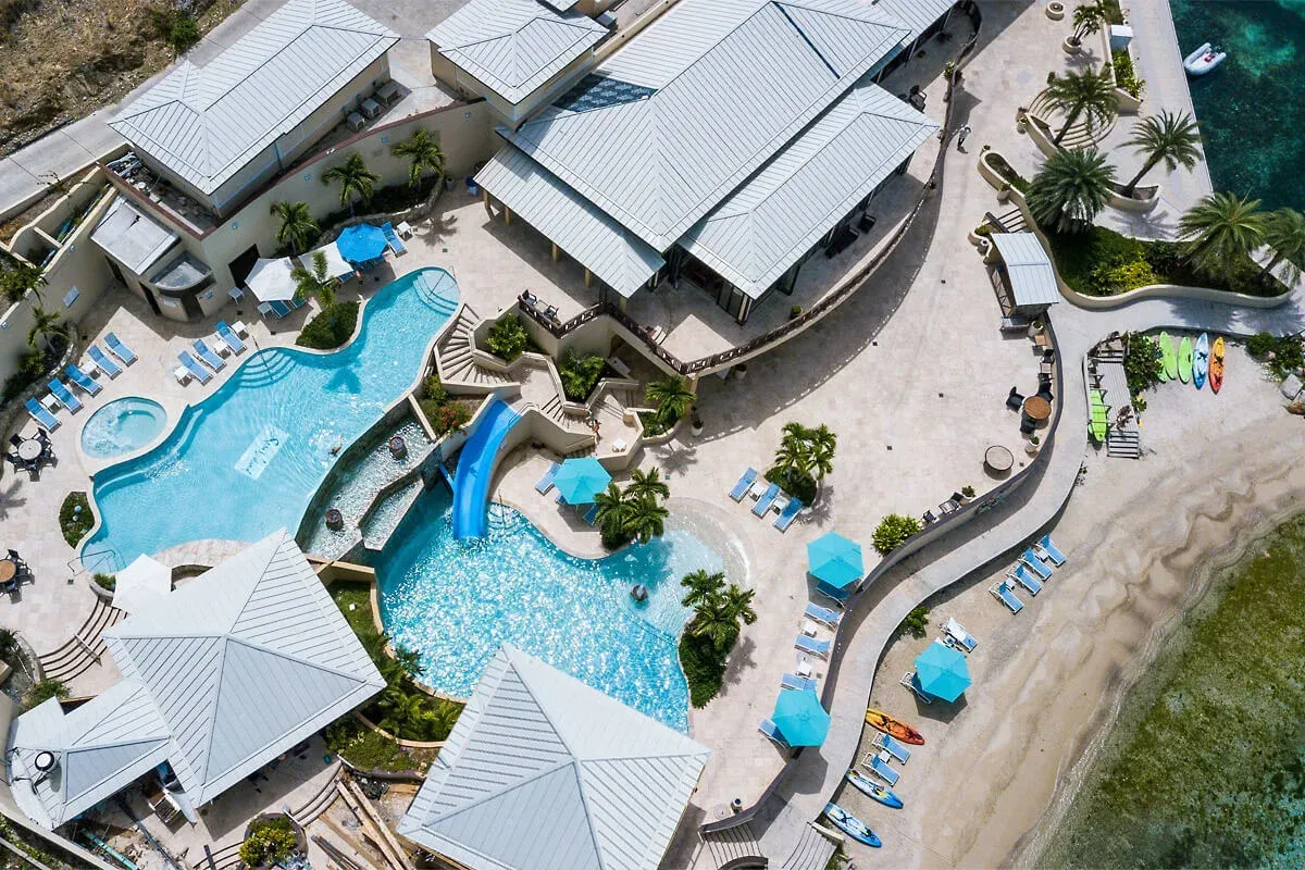 The pool and pavilions from directly above, a slide, blue parasols, kayaks on the sand