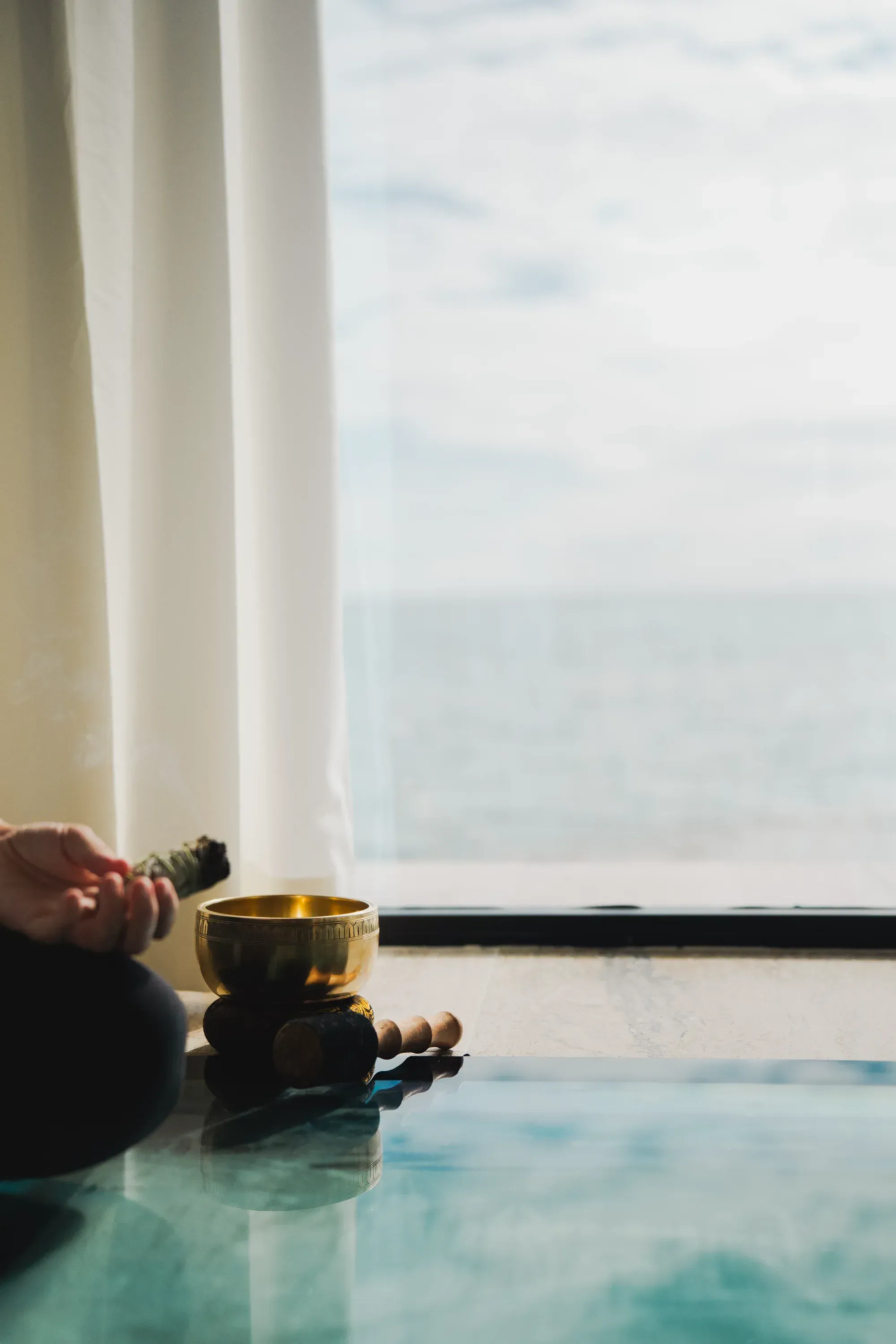 A hand holding smoking sage beside a brass singing bowl, the sea through the window