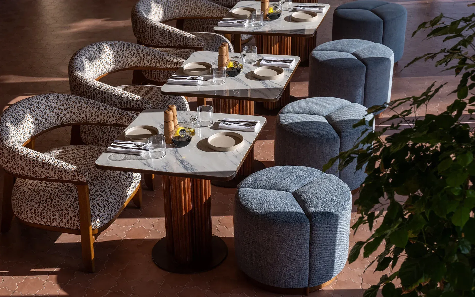 Marble tables set for lunch, patterned chairs and blue poufs on a terracotta floor