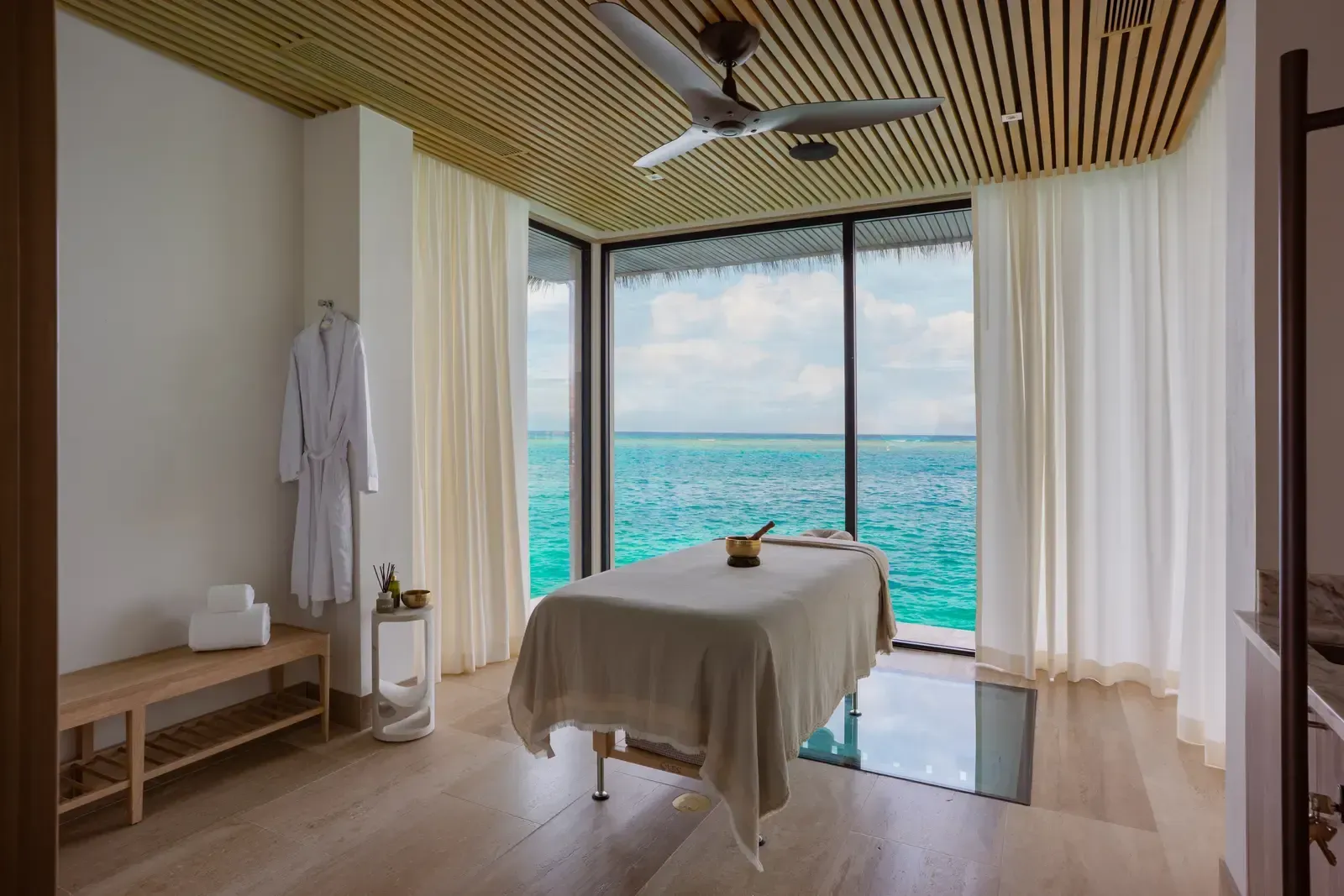 A treatment room: a slatted ceiling, white curtains, a glass floor, the sea through the corner
