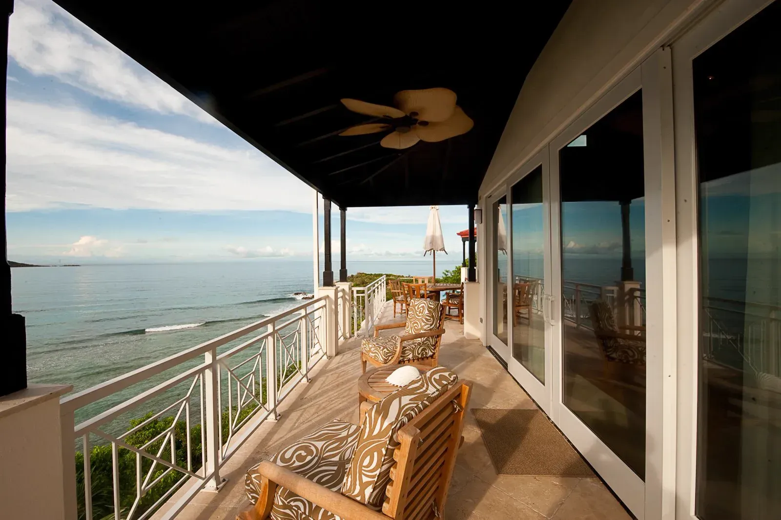 A villa balcony under a dark ceiling, armchairs facing the sea, a fan turning